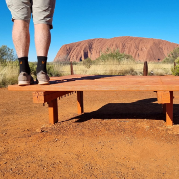 Close-up of legs wearing black U-branded socks standing on a timber platform with Uluru rising in the red desert behind them
